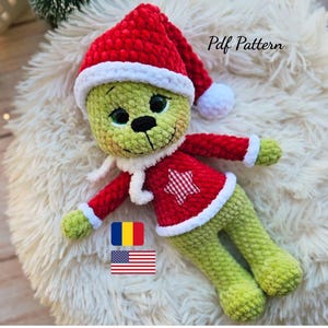 Puede incluir: Un personaje verde tejido a ganchillo con un gorro de Papá Noel rojo y un suéter rojo a juego con una estrella blanca. El personaje tiene una bufanda blanca y está acostado sobre una superficie blanca y esponjosa. El texto "Pdf Pattern" es visible.