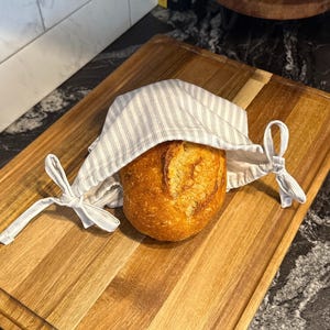 May include: A loaf of freshly baked bread rests on a wooden cutting board, partially covered by a striped cloth bread bag. The bread has a golden-brown crust. The cutting board has a rectangular shape.