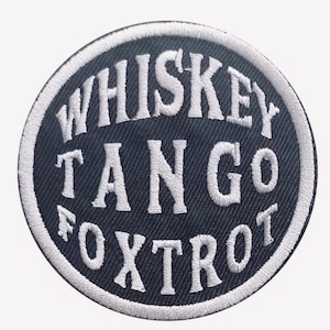 May include: A round, navy blue patch with white embroidered text. The text reads "WHISKEY TANGO FOXTROT" in a bold, sans-serif font. The patch has a white border and is likely designed to be sewn onto clothing or accessories.