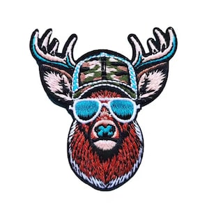 May include: Embroidered patch featuring a deer head wearing sunglasses and a camouflage hat. The deer has a red and brown face with blue sunglasses and a blue and green hat. The patch has a black border.