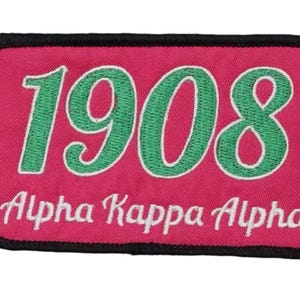 May include: A rectangular patch with a pink background and black border. The patch features the embroidered text "1908" in green with a white outline, and "Alpha Kappa Alpha" in white.