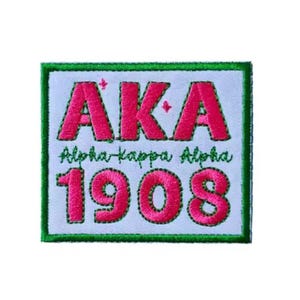 May include: Embroidered patch with the letters "AKA" in pink, above "Alpha Kappa Alpha" in script, and the number "1908". The patch has a white background with a green border.