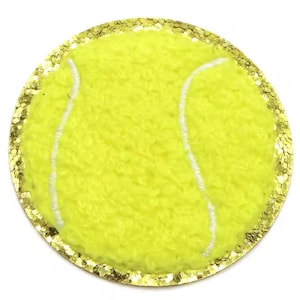 May include: A round, yellow tennis ball patch with white stitching and a gold glitter border. The patch has a fuzzy texture and is set against a white background. The design is simple and eye-catching.