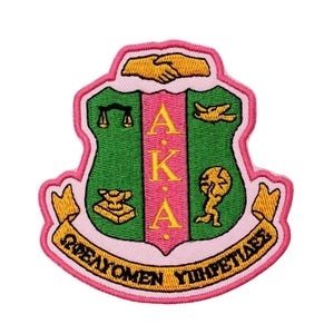May include: Embroidered patch featuring the letters AKA in yellow, set against a pink and green shield design. The patch includes various symbols and Greek text on a pink banner, all outlined in pink.