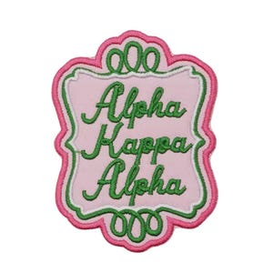 May include: Pink, green, and white embroidered patch with the text "Alpha Kappa Alpha". The patch has a decorative border and is designed with a pink background and green lettering.