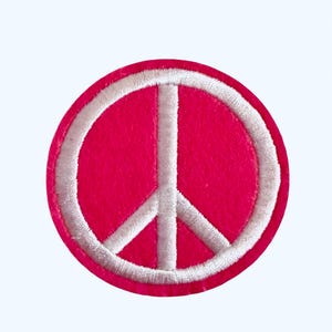 May include: A round, hot pink patch featuring a white peace symbol. The peace sign is a white, embroidered design. The patch is a circle shape, with a white embroidered border. This patch can be used to decorate clothing or accessories.