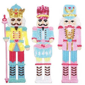 May include: Three embroidered nutcracker patches featuring sequin embellishments. The left figure wears a gold crown and holds a staff. The center figure plays a light blue drum. The right figure wears a light blue hat with a gold crescent moon.