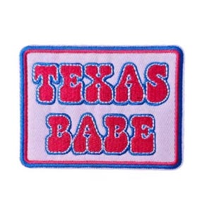 May include: A rectangular patch with the words "TEXAS BABE" in red, bubble letters. The text is outlined in white and set against a light pink background. The patch has a blue border.