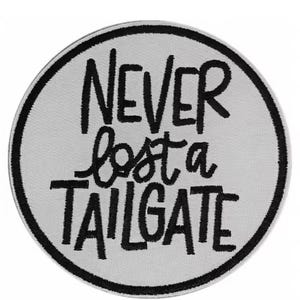 May include: Circular patch with the embroidered phrase "NEVER lost a TAILGATE" in black on a light gray background. The patch has a black border. The text is in a mix of block and cursive fonts.
