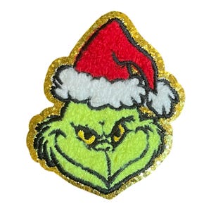 May include: A green, fuzzy Grinch face with a red Santa hat and white trim. The Grinch's face has black outlines and yellow eyes. The image is outlined with gold glitter.