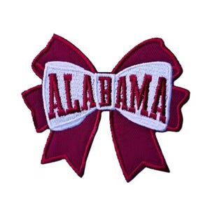 May include: A maroon and white embroidered bow-shaped patch. The patch features the word "ALABAMA" in maroon letters on a white background. The bow has a maroon border and is set against a white background.