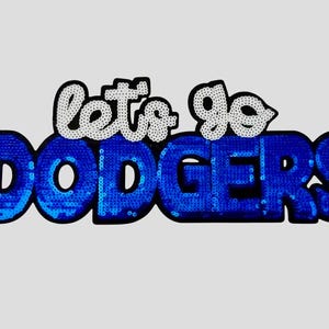 May include: A sequined patch with the words "let's go DODGERS" in a stylized font. The word "DODGERS" is in blue sequins, and "let's go" is in white sequins. The patch is on a light gray background.