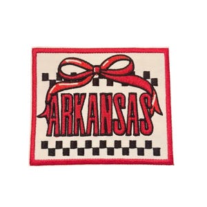 May include: Embroidered patch featuring the word "ARKANSAS" in red, with a red bow at the top. The patch has a cream-colored background with a black and white checkered pattern and a red border.