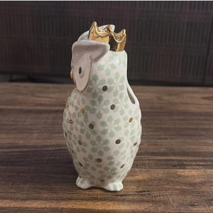 Może przedstawiać: Ceramiczna figurka sowy ze złotą koroną. Sowa jest biała z zielonym kwiatowym wzorem i złotymi akcentami. Urocza dekoracja do domu.