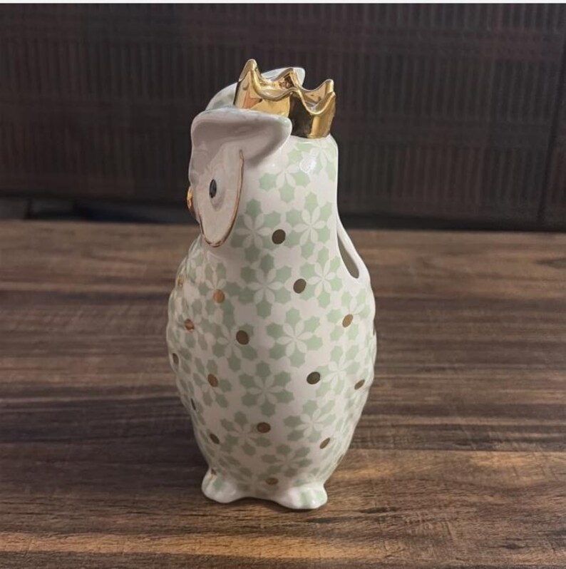 Może przedstawiać: Ceramiczna figurka sowy ze złotą koroną. Sowa jest biała z zielonym kwiatowym wzorem i złotymi akcentami. Urocza dekoracja do domu.