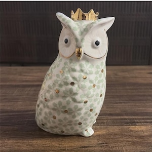 Może przedstawiać: Ceramiczna figurka sowy ze złotą koroną. Sowa ma biały i jasnozielony kwiatowy wz&oacute;r ze złotymi akcentami. Ma czarne oczy i złote detale. Dekoracyjny przedmiot do domu.