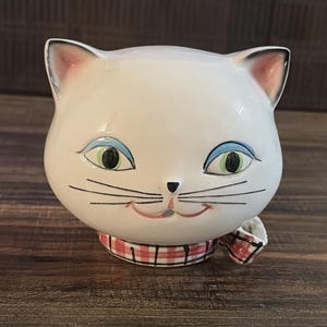 Puede incluir: Un recipiente de cerámica blanca con forma de gato con una cara sonriente, sombra de ojos azul y ojos verdes. El gato tiene una bufanda a cuadros rojos y blancos. Probablemente un artículo decorativo o un pequeño recipiente de almacenamiento.