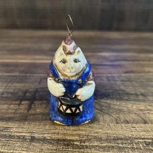 May include: A ceramic ornament of a cat wearing overalls and holding a drum. The cat is white with brown accents and blue overalls. The drum is black with white stripes.