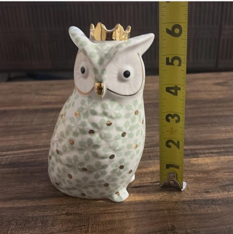 Może przedstawiać: Ceramiczna figurka sowy, o wysokości około 10 cm, ze złotą koroną. Sowa jest biała z jasnozielonym kwiatowym wzorem i złotymi akcentami. Ma czarne oczy i stoi wyprostowana.
