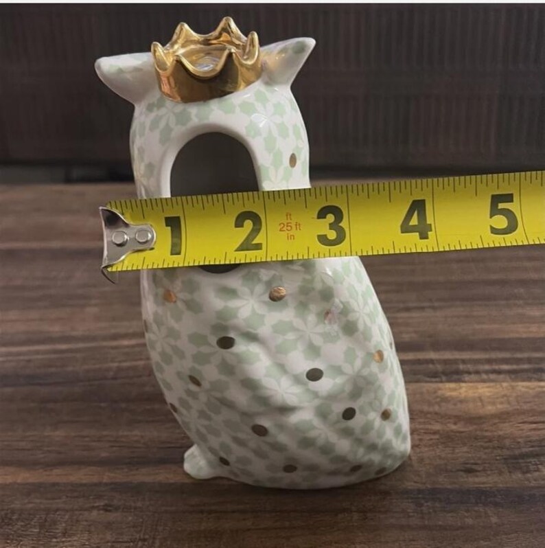Może przedstawiać: Ceramiczna figurka sowy ze złotą koroną, o wysokości około 10 cm. Sowa jest biała z jasnozielonym kwiatowym wzorem i złotymi akcentami. Sowa ma duży otw&oacute;r pośrodku.
