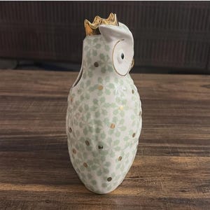 Może przedstawiać: Ceramiczna figurka sowy ze złotą koroną. Sowa jest biała z jasnozielonym kwiatowym wzorem i złotymi kropkami. Figurka ma duże, okrągłe oczy i mały dzi&oacute;b.