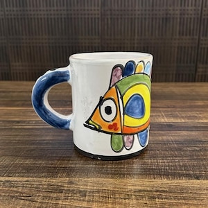 May include: White ceramic mug with a blue handle and a colorful cartoon fish design. The fish is yellow, orange, green, blue, and purple.