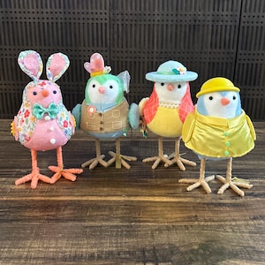May include: Four handmade Easter bird figurines. The birds are made of fabric and have different colors and patterns. One bird is wearing a pink bunny costume, another is wearing a blue and green jacket, another is wearing a yellow dress and hat, and the last one is wearing a yellow raincoat.