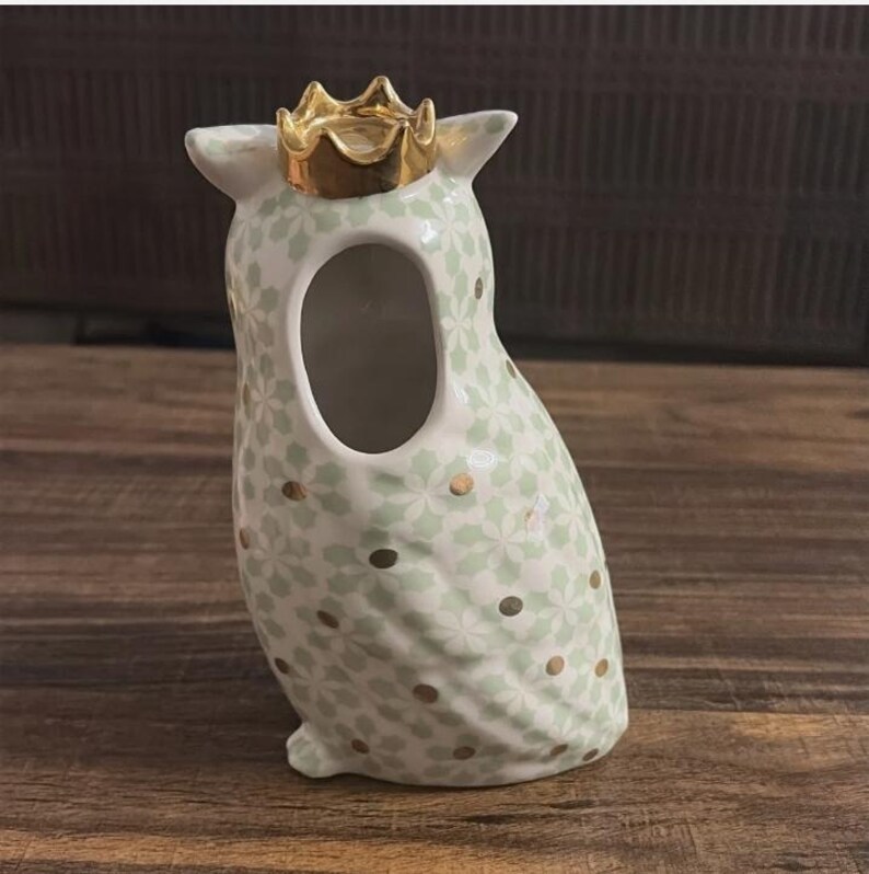 Może przedstawiać: Ceramiczny wazon w kształcie sowy ze złotą koroną. Wazon jest biały z jasnozielonym kwiatowym wzorem i złotymi kropkami. Sowa ma duży otw&oacute;r pośrodku.