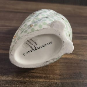 Może przedstawiać: Ceramiczny przedmiot dekoracyjny w kształcie sowy z jasnozielonym kwiatowym wzorem i złotymi akcentami. Sp&oacute;d sowy zawiera napis "by ANTHROPOLOGIE" oraz "Made in China".