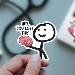 Pode incluir: Um adesivo com uma figura de pau de desenho animado segurando um cérebro rosa. O texto no adesivo diz "Hey, você perdeu isso".