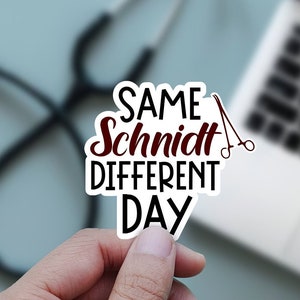 Pode incluir: Um adesivo branco com o texto "SAME Schmidt DIFFERENT DAY" em letras pretas e vermelhas. Um pequeno par de tesouras está ao lado do texto. O adesivo está sendo segurado por uma mão.