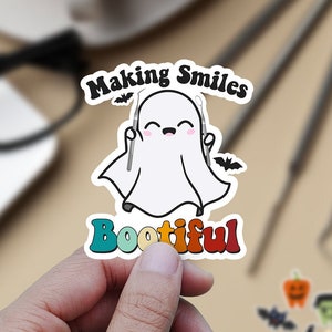 May include: A white ghost with a toothy grin holds two dental tools in its hands. The ghost is surrounded by a colorful rainbow-like border with the text "Making Smiles Bootiful" in a retro font.