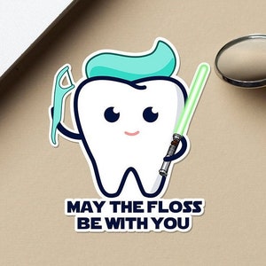 May include: A white cartoon tooth with a blue and green toothbrush and a green lightsaber. The text "May the floss be with you" is below the tooth.