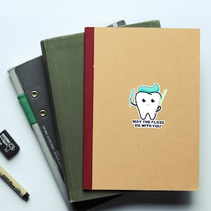May include: A brown notebook with a white sticker of a cartoon tooth holding a toothbrush and a lightsaber. The text on the sticker reads "May the floss be with you."