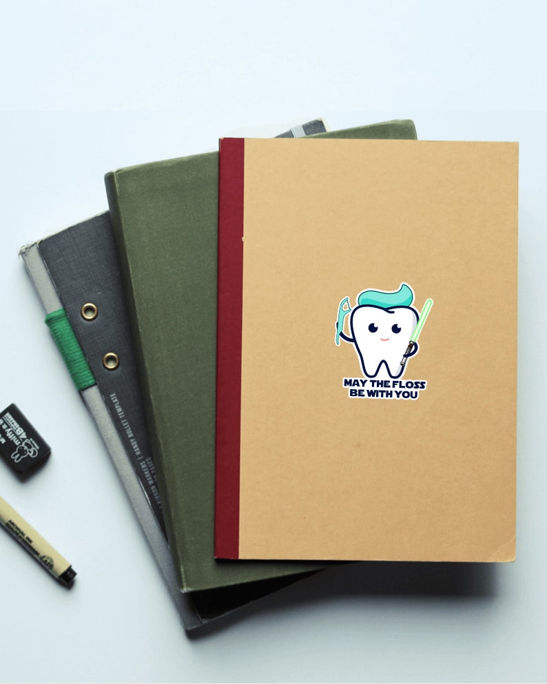 May include: A brown notebook with a white sticker of a cartoon tooth holding a toothbrush and a lightsaber. The text on the sticker reads "May the floss be with you."
