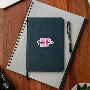 Puede incluir: Una libreta azul oscuro con una pegatina rosa y negra que dice "NP Your Name" en la portada. La libreta est&aacute; sobre una superficie de madera con un bol&iacute;grafo y una planta.