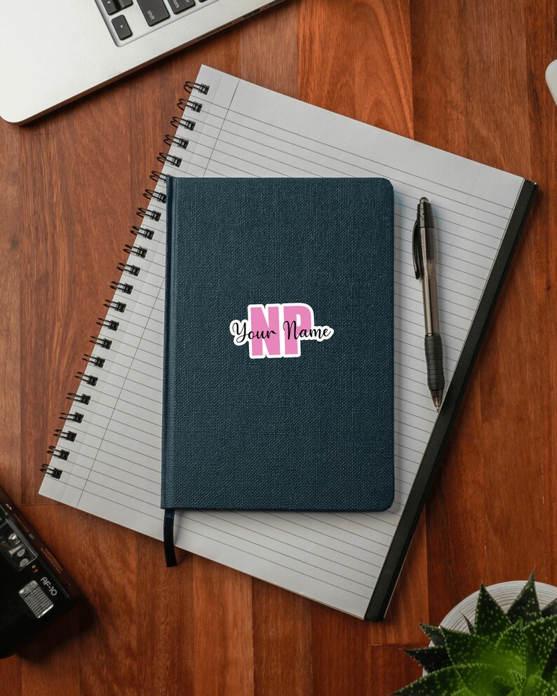 Puede incluir: Una libreta azul oscuro con una pegatina rosa y negra que dice "NP Your Name" en la portada. La libreta est&aacute; sobre una superficie de madera con un bol&iacute;grafo y una planta.