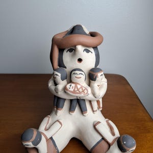 May include: A hand-painted clay storyteller figurine featuring a large central figure with a brown snake draped over its head, holding three smaller figures in its arms, and two additional small figures resting on its outstretched legs.