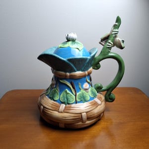May include: A ceramic teapot featuring a blue body with green leaf motifs and a tan woven basket-style base. The lid is topped with a white flower, and the green handle is shaped like a branch with a small green frog perched on the top.