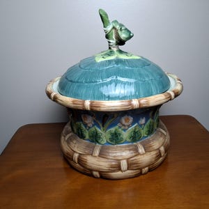 May include: A ceramic lidded tureen featuring a base designed to resemble a woven brown basket. The middle section displays a blue band with painted pink flowers and green leaves. The lid is teal with a sculpted green leaf handle on top.