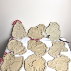 May include: A collection of eight beige ceramic cookie molds featuring various holiday designs including a Christmas tree, rocking horse, angel, Santa Claus, and a wreath, each with a small hole and a red ribbon for hanging.