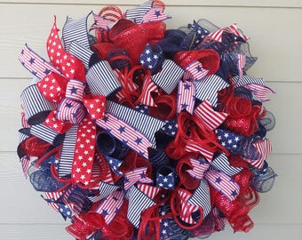 May include: A red, white, and blue patriotic wreath with a mesh base and ribbon accents. The ribbon features a white and blue striped pattern with red stars.
