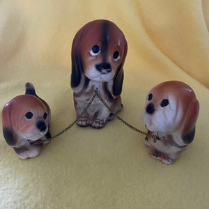 May include: Three ceramic dog figurines, two brown and white and one brown and black, are connected by gold chains. The dogs are all looking down with sad expressions.