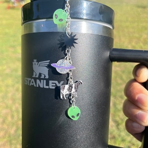 May include: Black stainless steel travel mug with a black and white cow, a planet, a star, and two green aliens hanging from a silver chain attached to the lid.