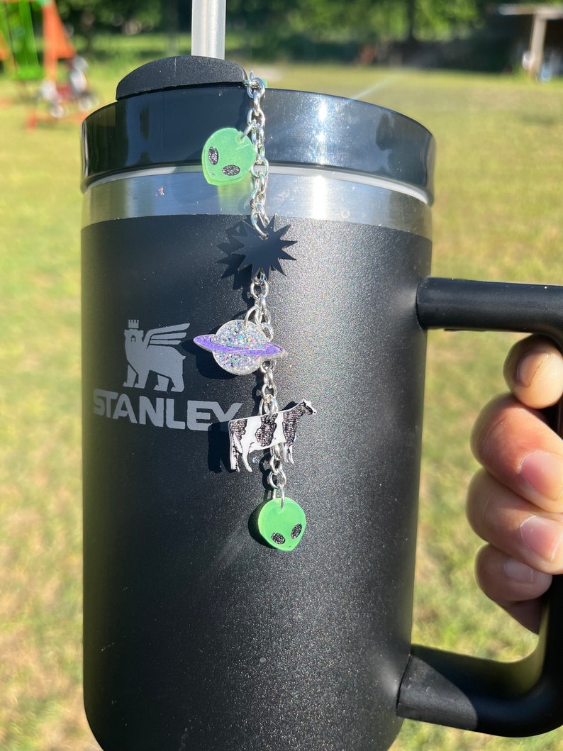 May include: Black stainless steel travel mug with a black and white cow, a planet, a star, and two green aliens hanging from a silver chain attached to the lid.