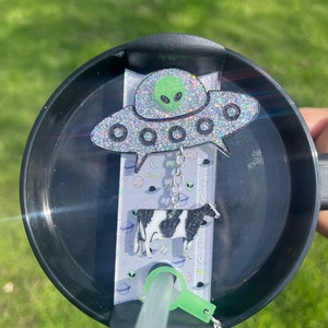 May include: A black and white cow charm hangs from a silver chain attached to a green glitter UFO charm. The charms are attached to a black lid with a straw holder. The lid is on a clear plastic straw.