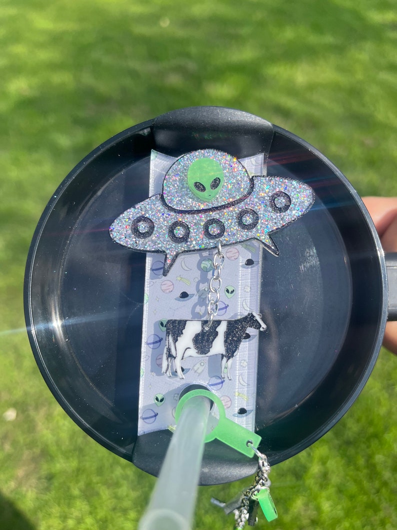 May include: A black and white cow charm hangs from a silver chain attached to a green glitter UFO charm. The charms are attached to a black lid with a straw holder. The lid is on a clear plastic straw.