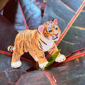 May include: A small, orange and black tiger cub figurine with a white stripe on its forehead, standing on a dark green leaf with red veins.