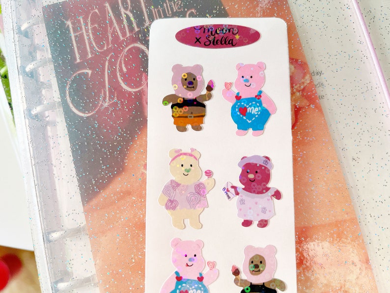 May include: A sheet of stickers featuring cute cartoon bears with different expressions and outfits. The stickers are designed with a pastel color scheme and feature the text "me x Stella".