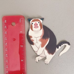May include: A white, brown, and black cartoon animal with a long tail. The animal has a white face with a pink nose and a wide smile. The animal is about 3 inches tall.